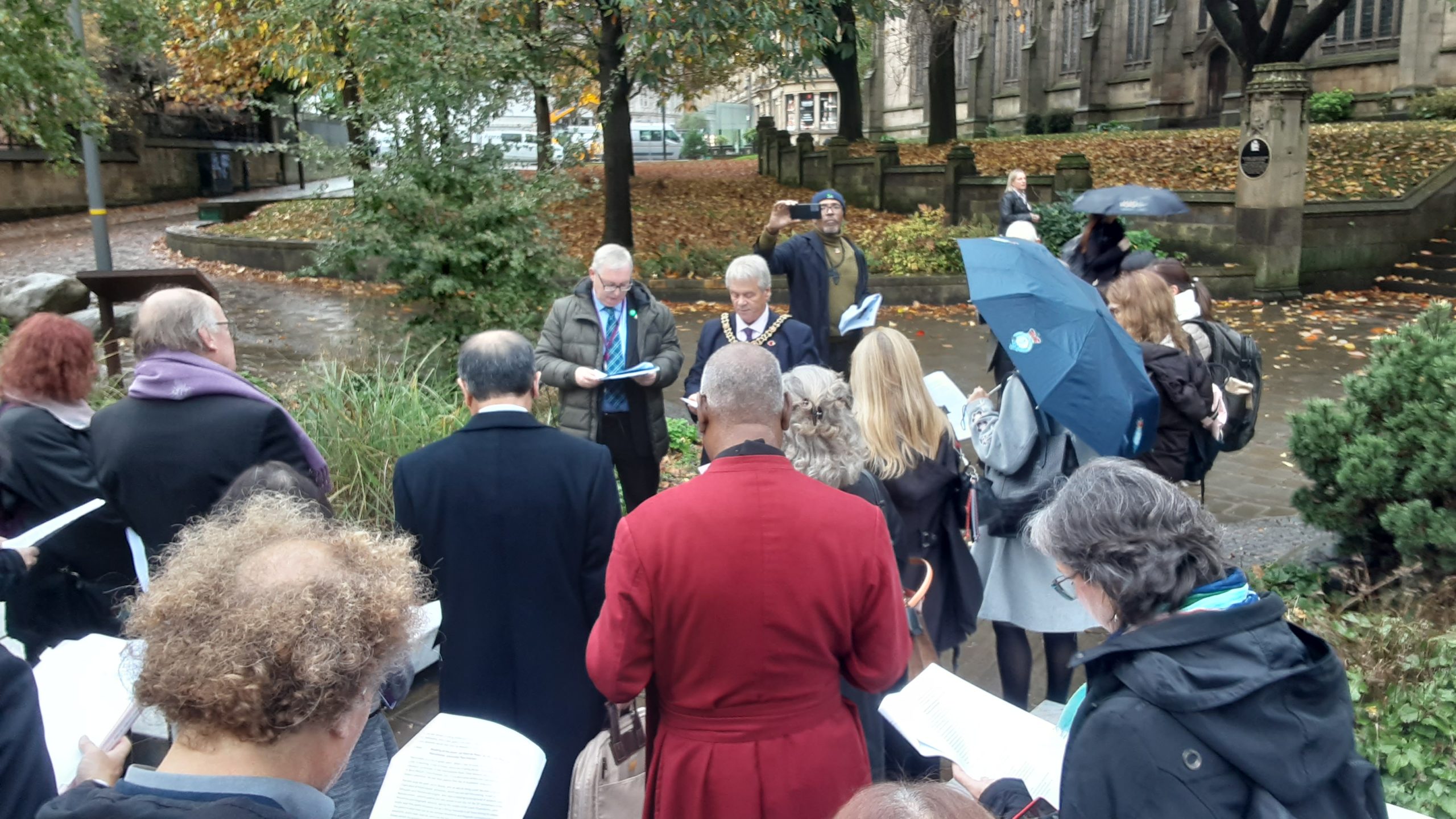 Group of people standing outside facing a mayor, reading from sheets of paper. It is raining